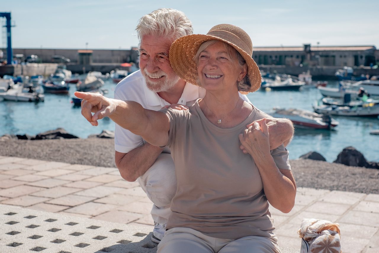 Smiling senior couple sitting together on a bench at a sunny harbor